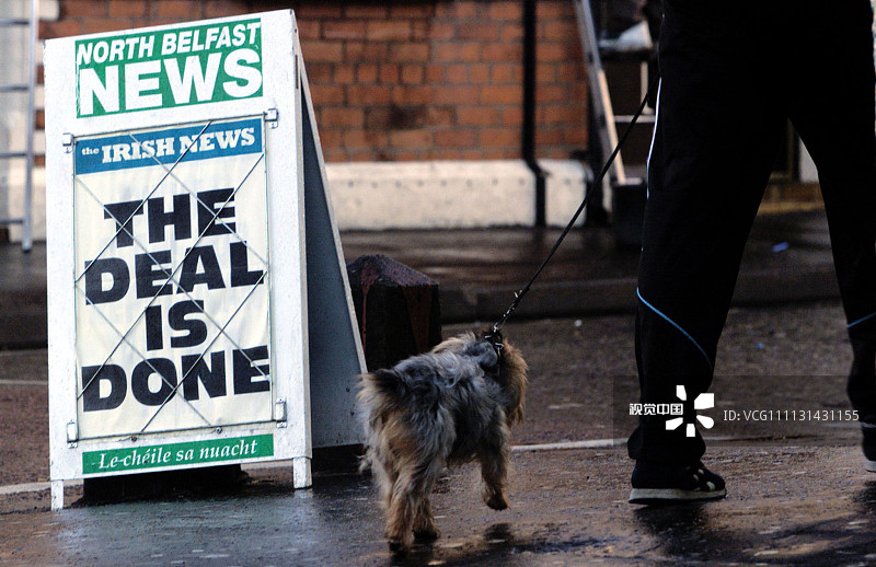 a man walks his dog past a newspaper bill board in belfast.